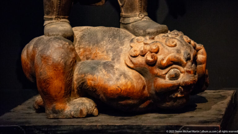 Close-up of a muscular carved demon lying beneath the feet of a standing guardian.