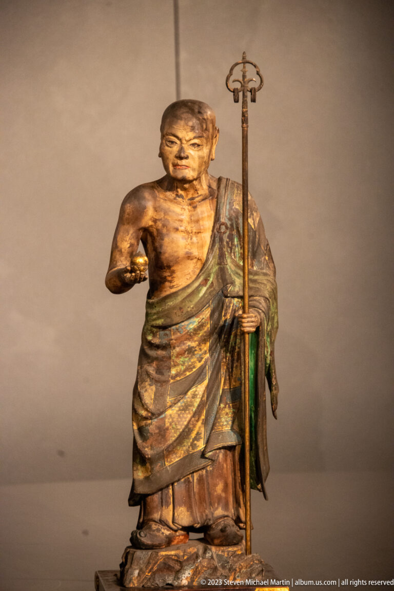 Standing wooden Buddhist monk holding a tall ringed staff and extending one hand.