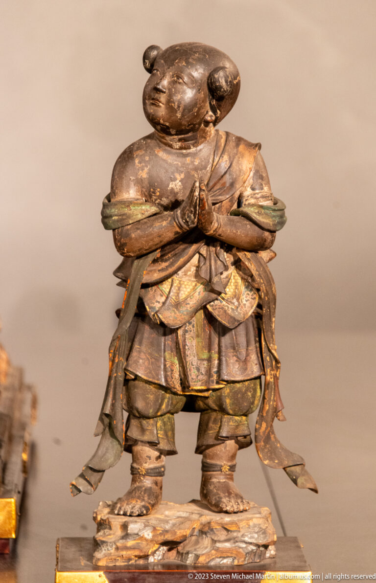 Animal-headed wooden guardian standing with hands joined in prayer and wearing layered robes.