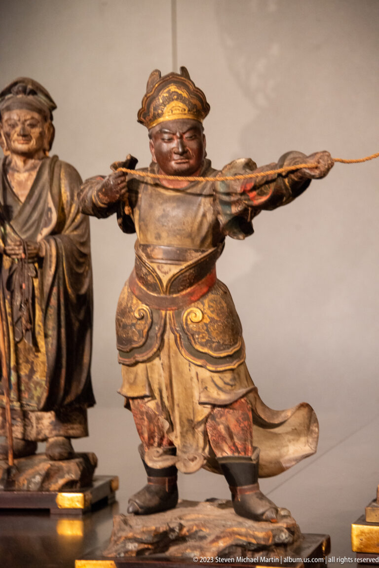 Polychrome wooden warrior in layered armor drawing a bow with arms extended.