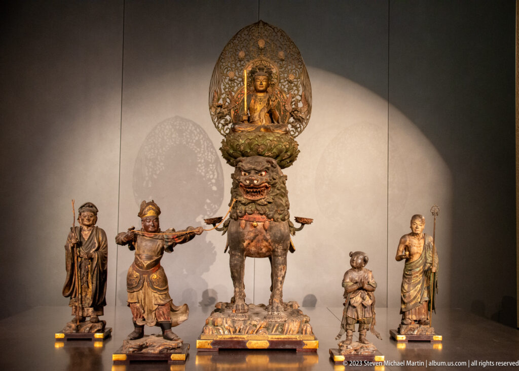 Five-figure Buddhist ensemble with central guardian, seated Buddha in mandorla, and flanking attendants.