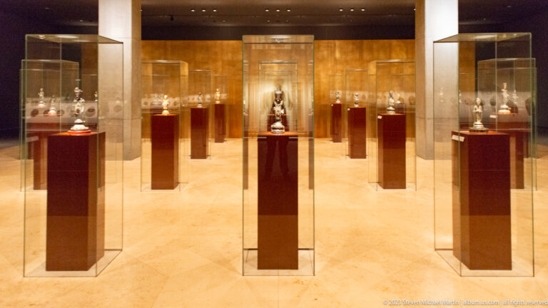 Warm museum gallery with small Buddhist statues in individual glass cases on dark red plinths.
