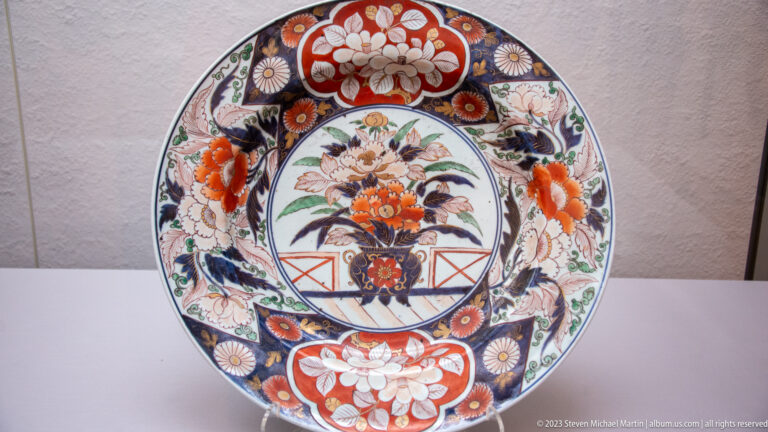 Large Japanese porcelain plate with central flower vase and red, blue, green, and gold border panels.