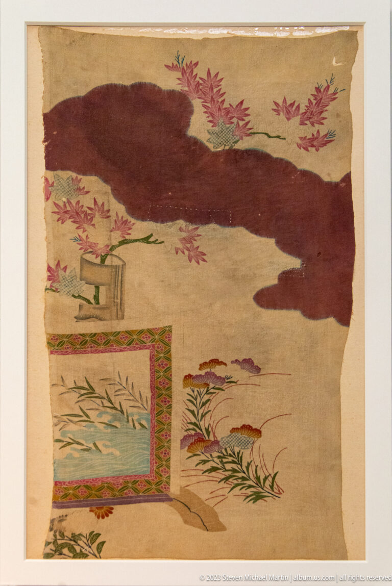 Fragmentary beige textile with dark tree form, red leaves, small architecture, and patterned borders.