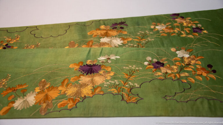 Green textile panel embroidered with white, orange, purple, and gold flowers and cloud-like bands.