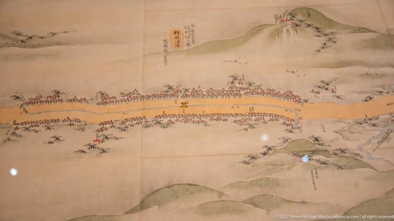 Painted procession crossing a bridge through a river valley with rounded mountains and many small figures.