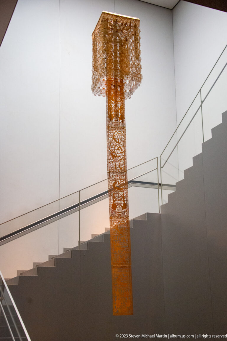 Long suspended openwork metal ornament with a square canopy hanging beside a modern museum stair.