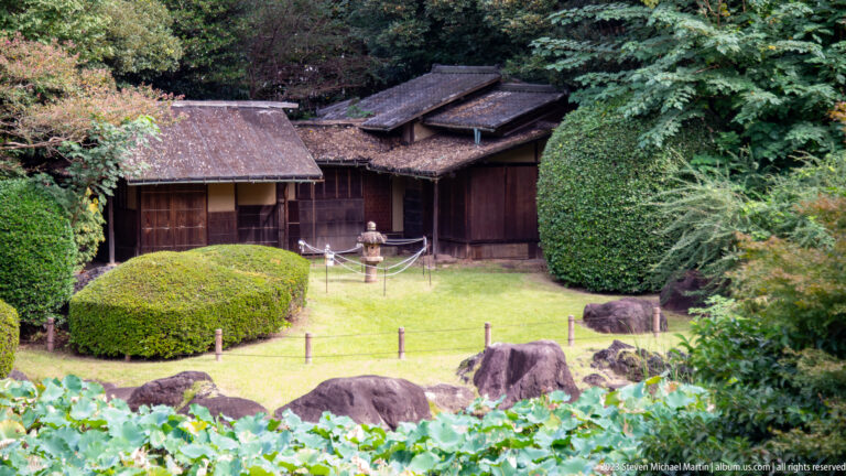 Rustic wooden garden building surrounded by clipped shrubs, rocks, and dense greenery.
