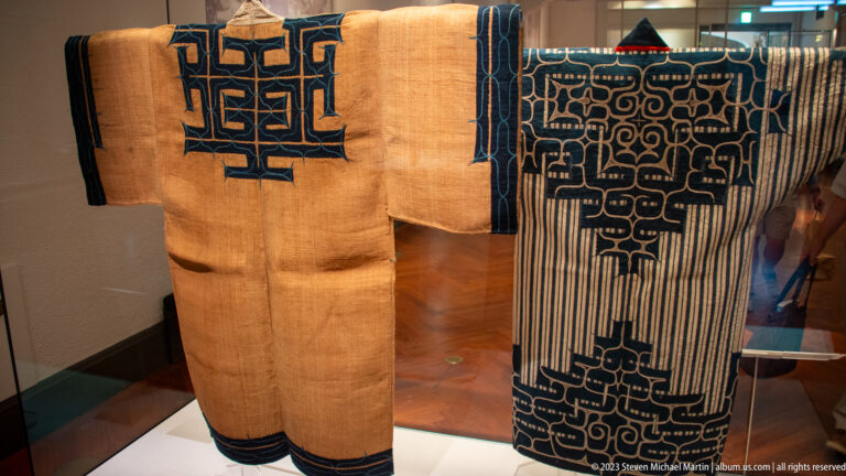Two natural-colored robes with bold dark-blue geometric appliqué and embroidery.