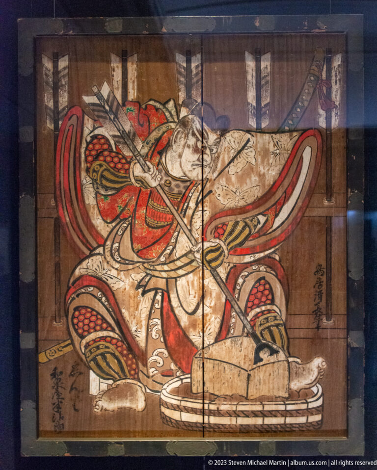 Painted armored warrior in red, black, and gold, crouching while holding a long weapon.