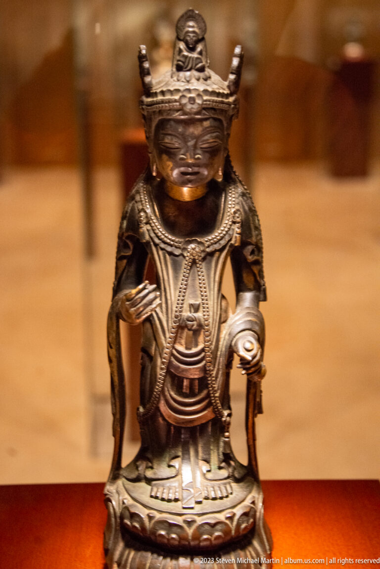 Small crowned gilt-bronze Buddhist figure standing frontally and holding objects in both hands.