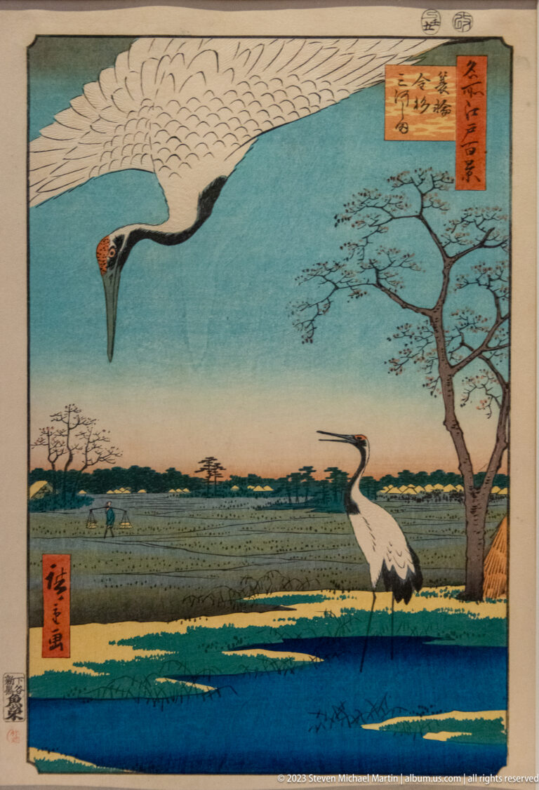 Japanese woodblock print with one crane flying overhead and another standing in a marsh.