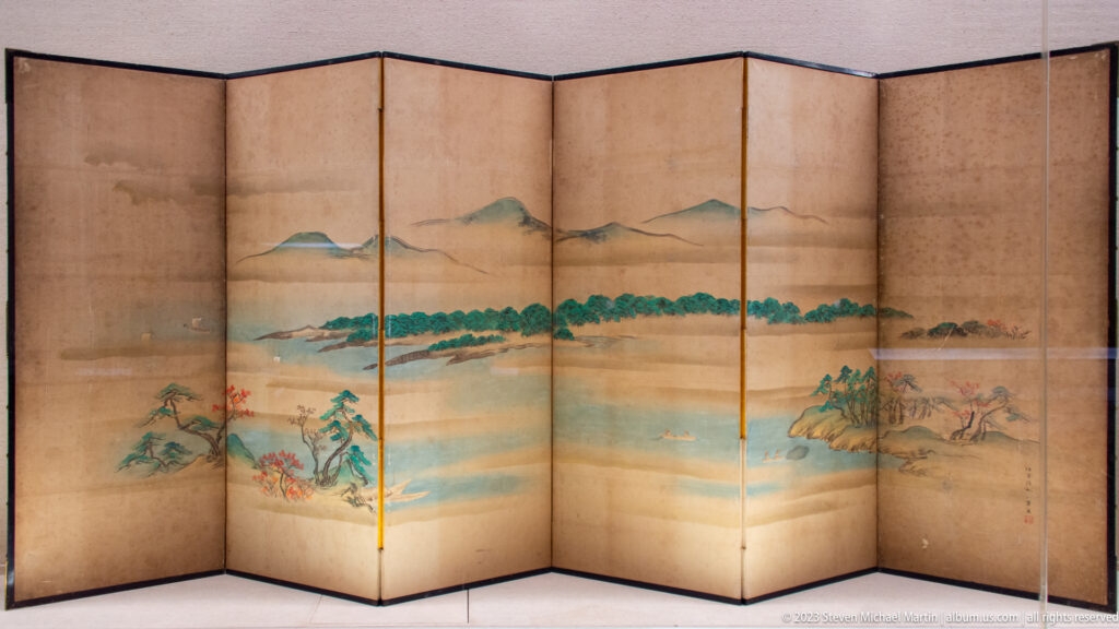Folding screen painted with water, distant mountains, wooded shores, and small landscape details.