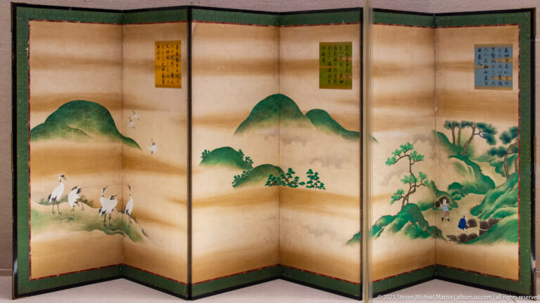Folding screen with green mountains, cranes, pine trees, figures, and calligraphy panels.