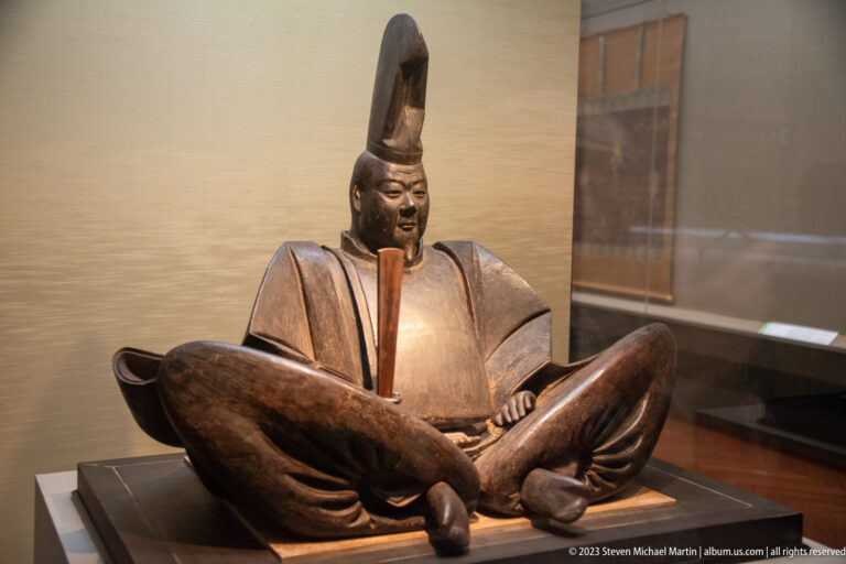 Seated wooden court figure wearing a tall black cap and formal angular robes.
