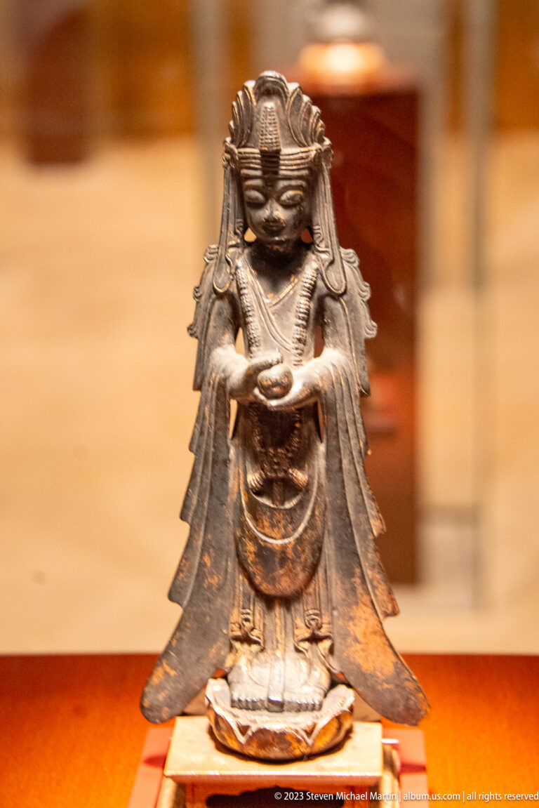 Small gilt-bronze Buddhist figure with tall headdress, gathered hands, and a flaring mantle.