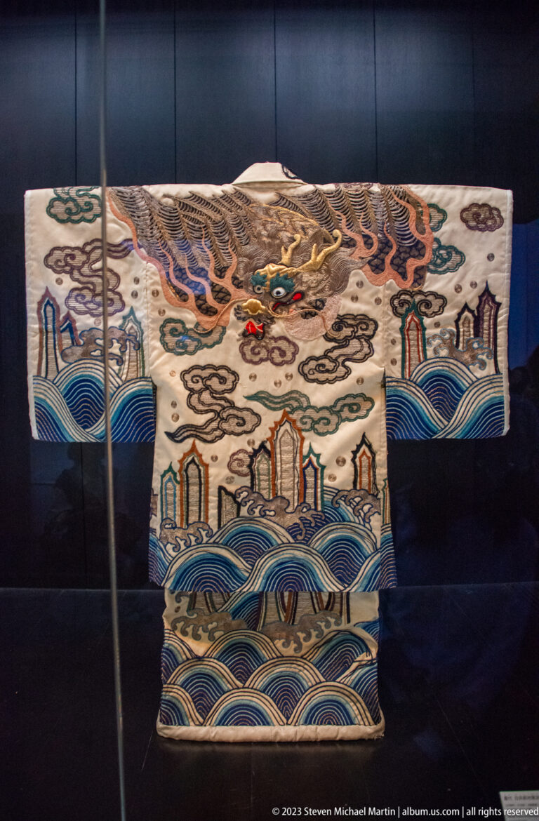 White theatrical robe embroidered with a dragon, clouds, blue waves, and architectural motifs.