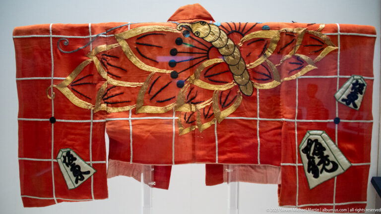 Red costume fragment with large gold butterfly, white grid lines, and Japanese character plaques.