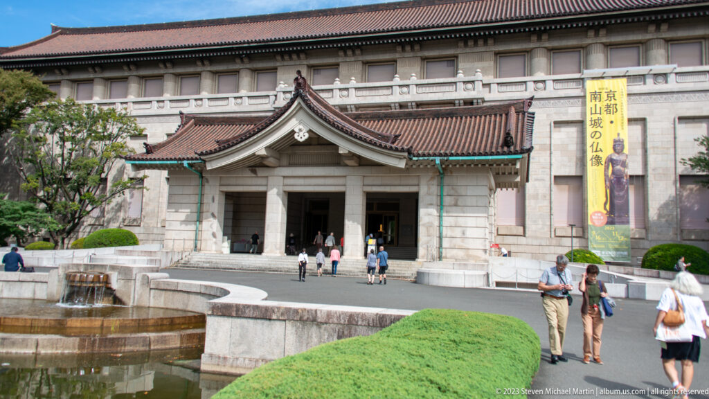 Honkan entrance with tiled roof, pale stone façade, visitors, fountain, and clipped shrubs.