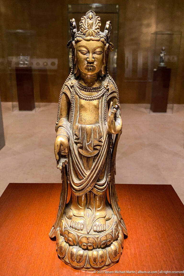 Small crowned gilt-bronze Buddhist figure standing on a lotus base with one palm open.
