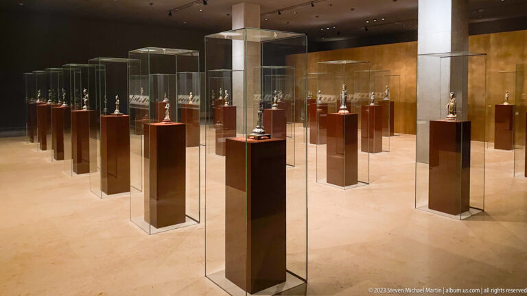 Warm museum gallery with rows of small Buddhist sculptures in individual glass cases on dark plinths.