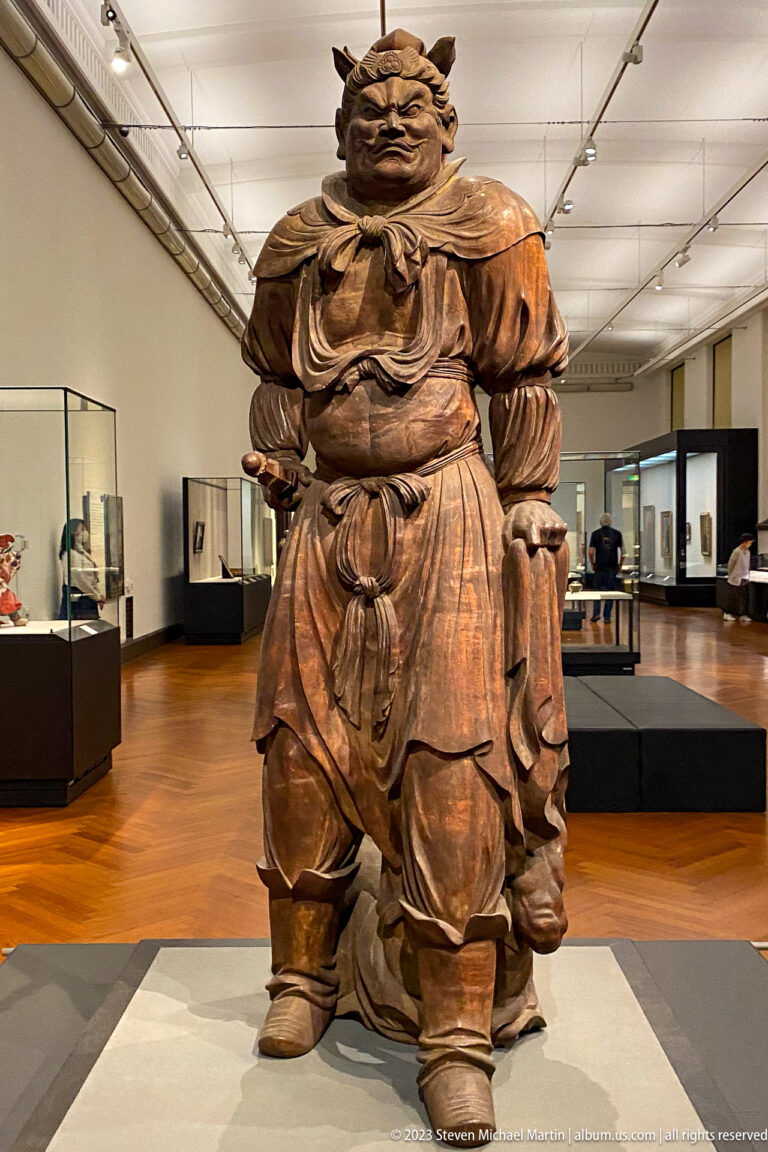 Large standing wooden Buddhist guardian with stern face, layered robes, and broad stance.