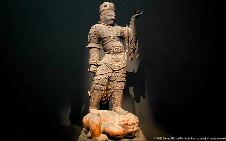 Armored standing Buddhist guardian on top of a prostrate demon-like figure.