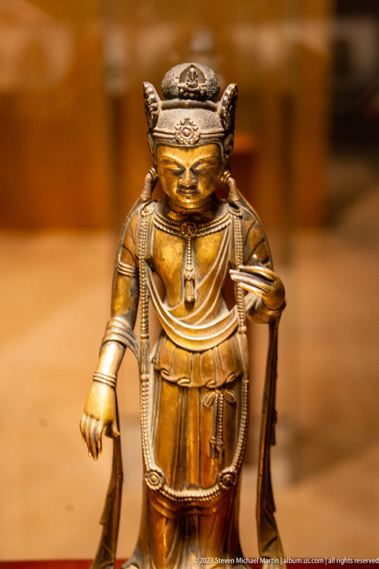 Golden crowned Buddhist figure standing with a long staff-like object and one hand lowered.