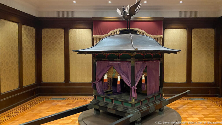 Compact imperial yōyo palanquin with long carrying poles, curved lacquered roof, purple curtains, and decorative finial.