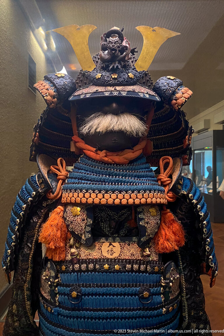 Front close-up of samurai armor with blue lacing, orange cords, fur trim, and large gold crest.