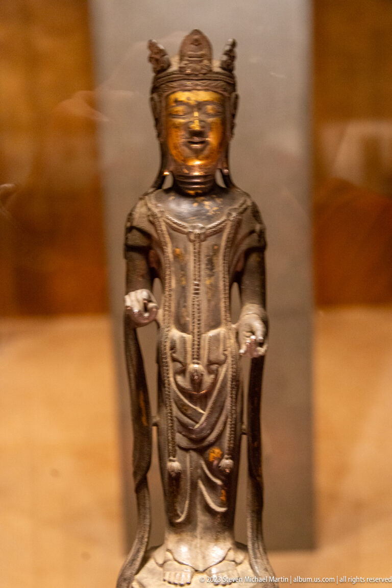 Dark crowned bronze Buddhist figure standing frontally with both hands extended, palms upward.