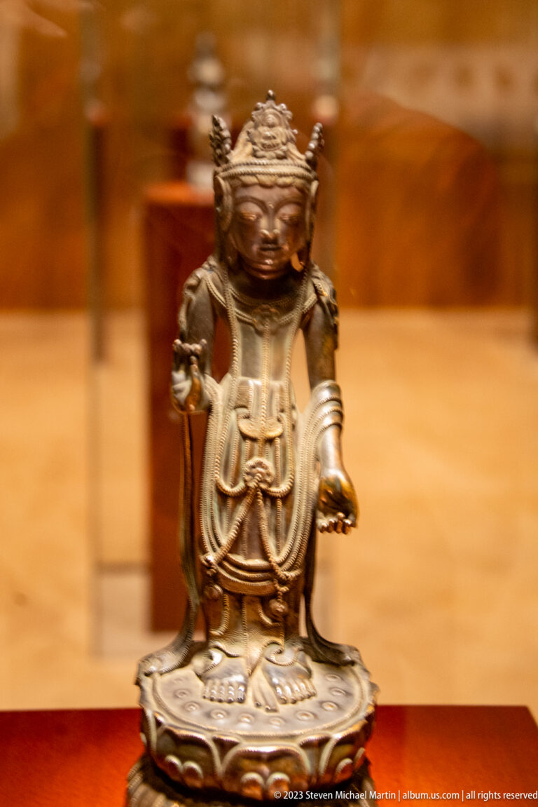Small dark gilt-bronze Buddhist figure on a lotus base with one hand raised and one extended.