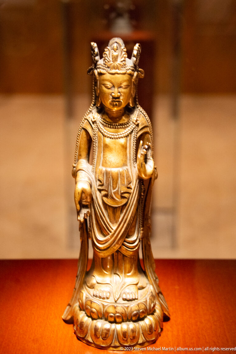 Small crowned golden Buddhist figure standing frontally and holding an object in each hand.