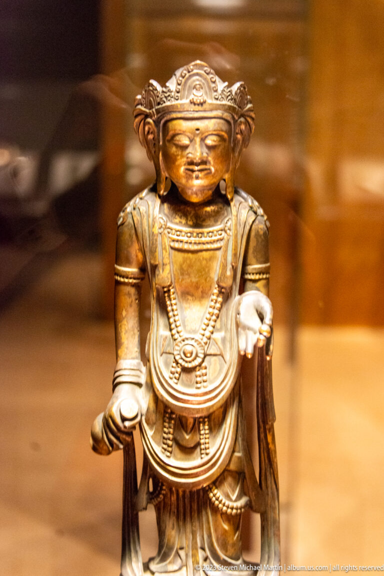 Small golden crowned Buddhist figure with one open palm extended and the other hand lowered.