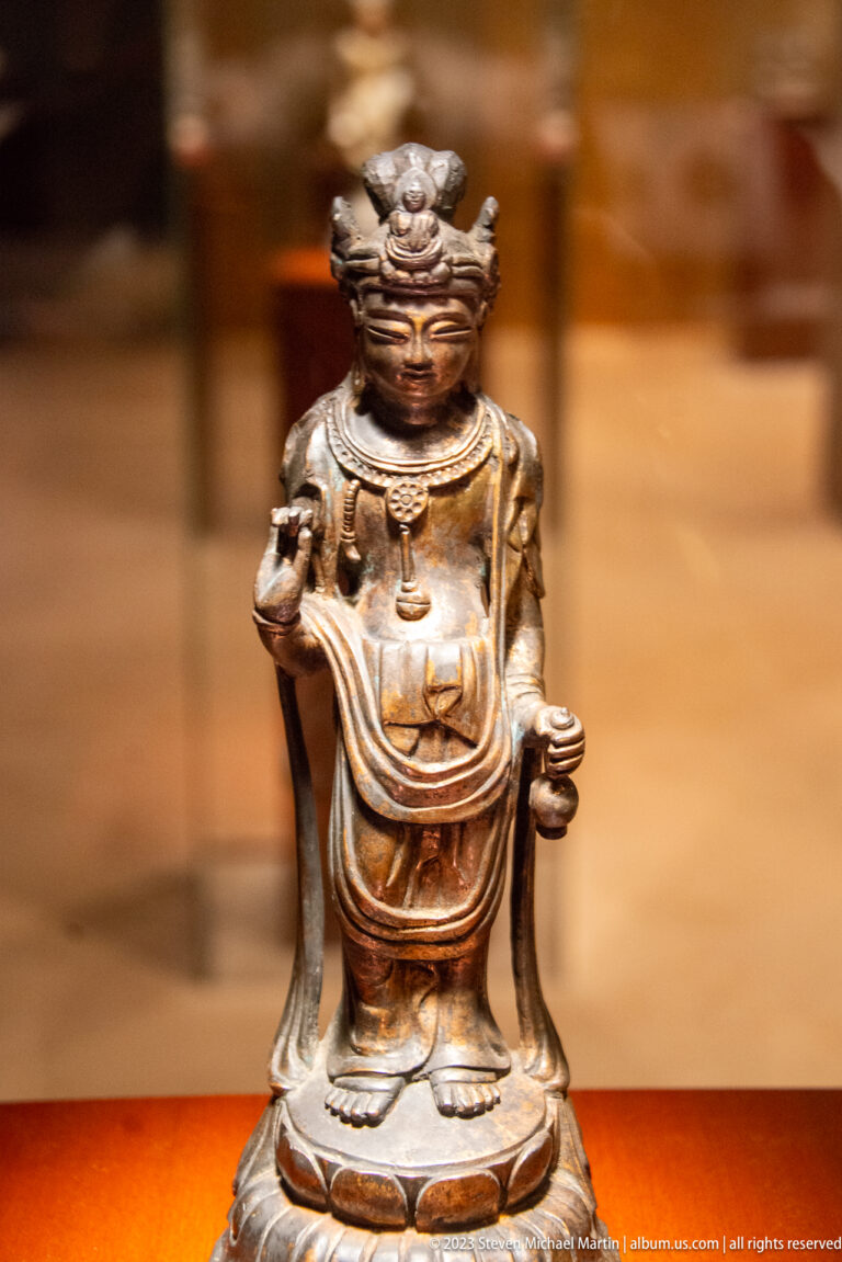 Small dark bronze Buddhist figure with one open hand raised and a small object in the other.