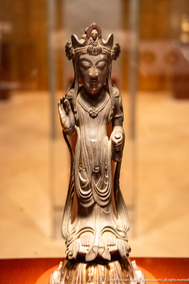 Small dark bronze Buddhist attendant on a lotus base with one hand raised and long flowing robes.