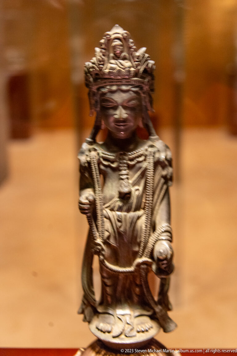 Small dark crowned Buddhist figure in a low seated posture with one hand extended.
