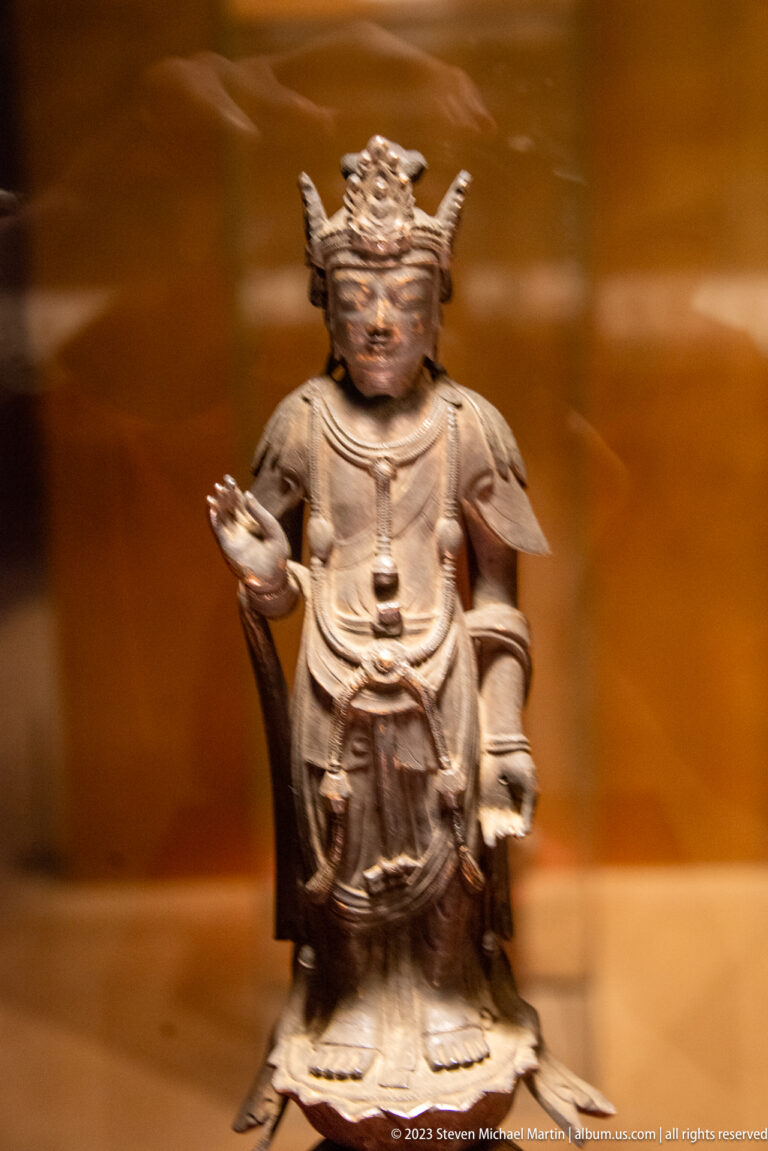 Small dark bronze guardian-like figure with one raised hand, broad shoulder drapery, and chest pendant.
