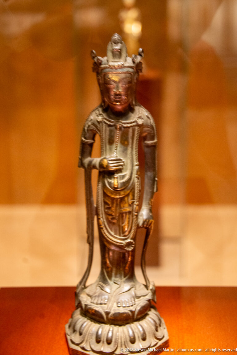Small crowned bronze Buddhist figure holding a narrow object across one hand.