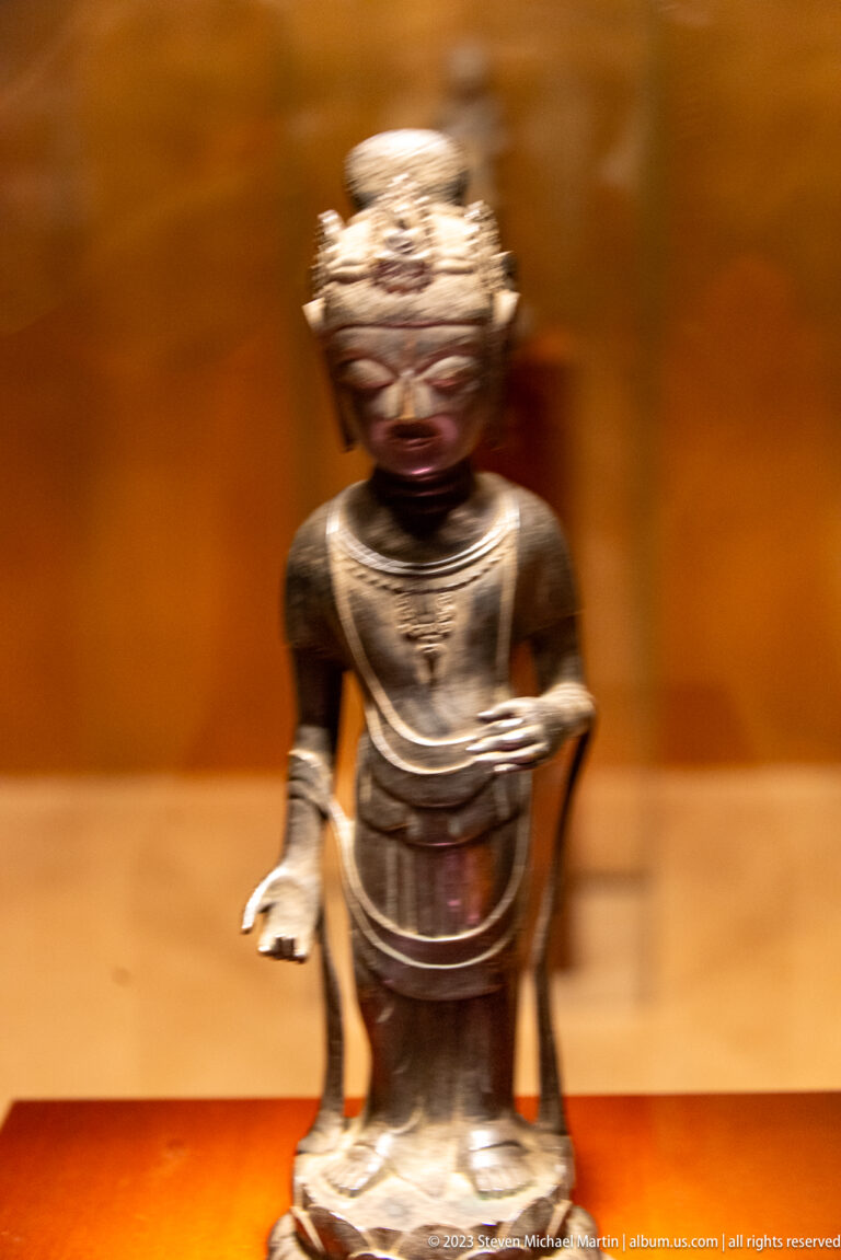Small dark bronze Buddhist figure leaning forward with one hand extended and the other lowered.