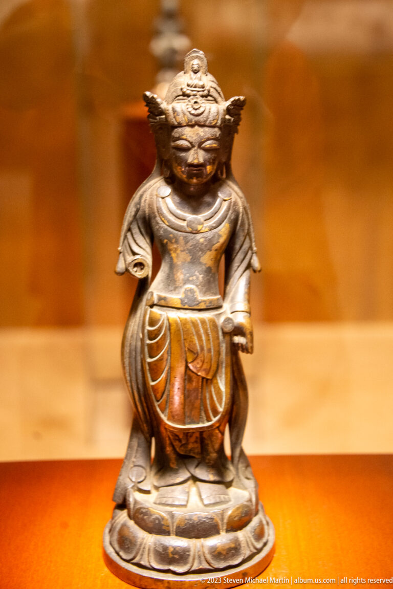 Small dark bronze guardian-like figure seated on a rounded base and holding a long object across the body.