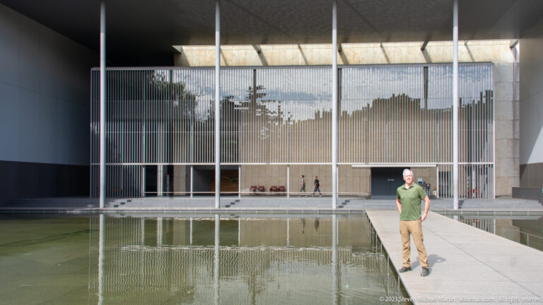 Man standing on a narrow walkway across a reflecting pool before the screened façade of the Horyuji gallery.
