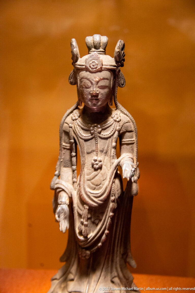 Small crowned bronze Buddhist figure standing with both hands open and layered drapery.