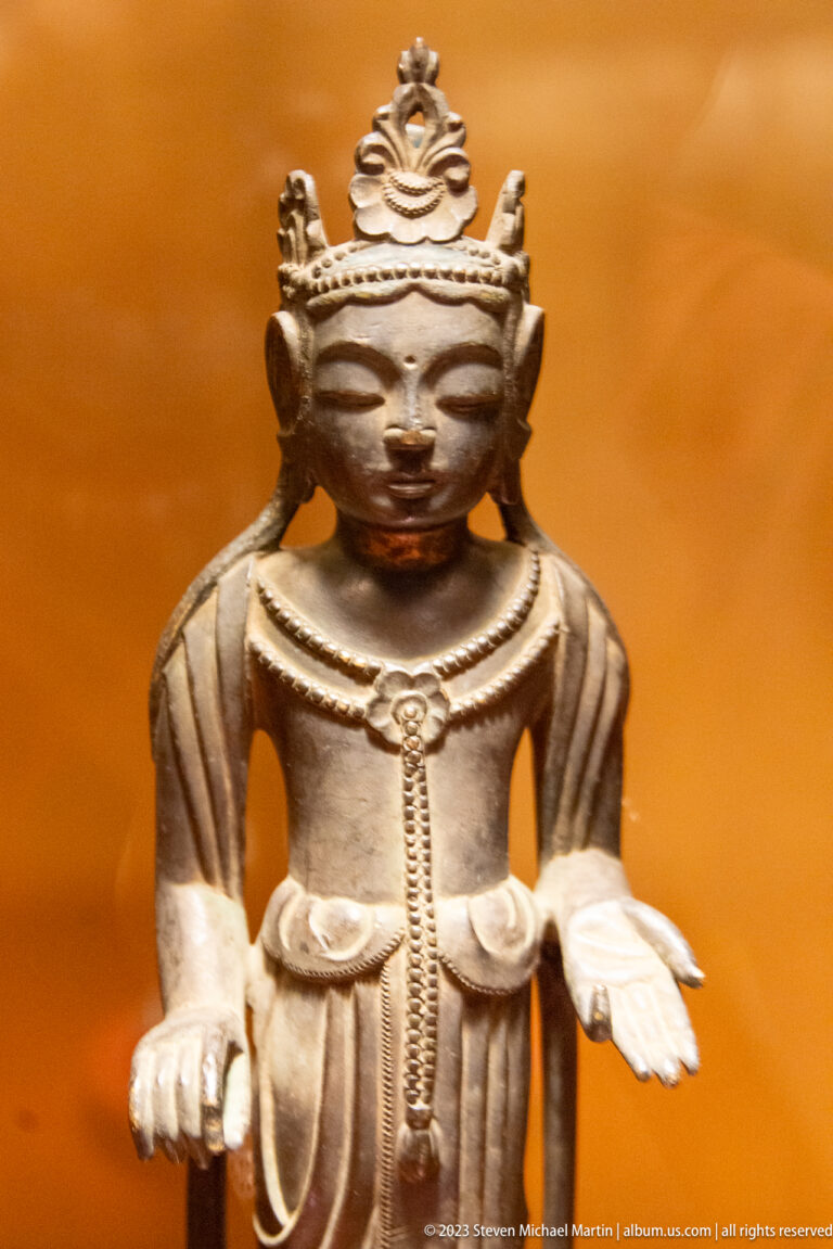 Close-up of crowned bronze Buddhist figure with both hands extended and a broad headdress.