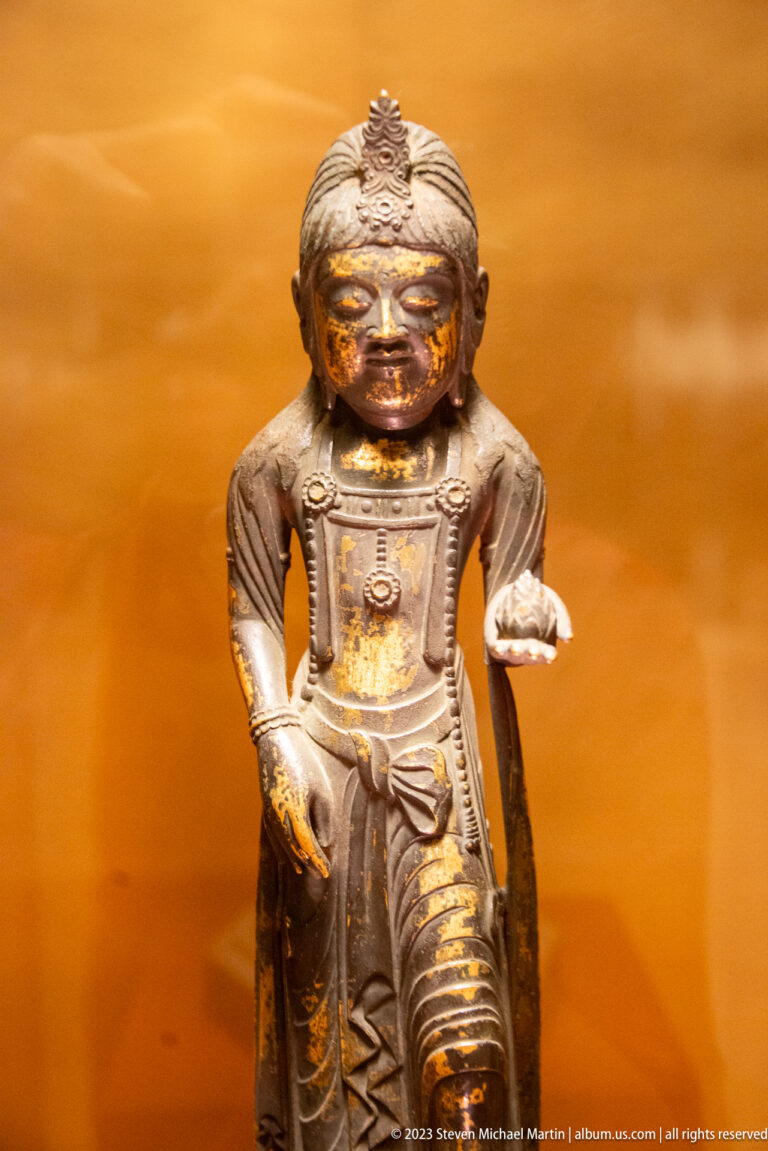 Small crowned bronze figure with one bent knee and one palm extended forward.