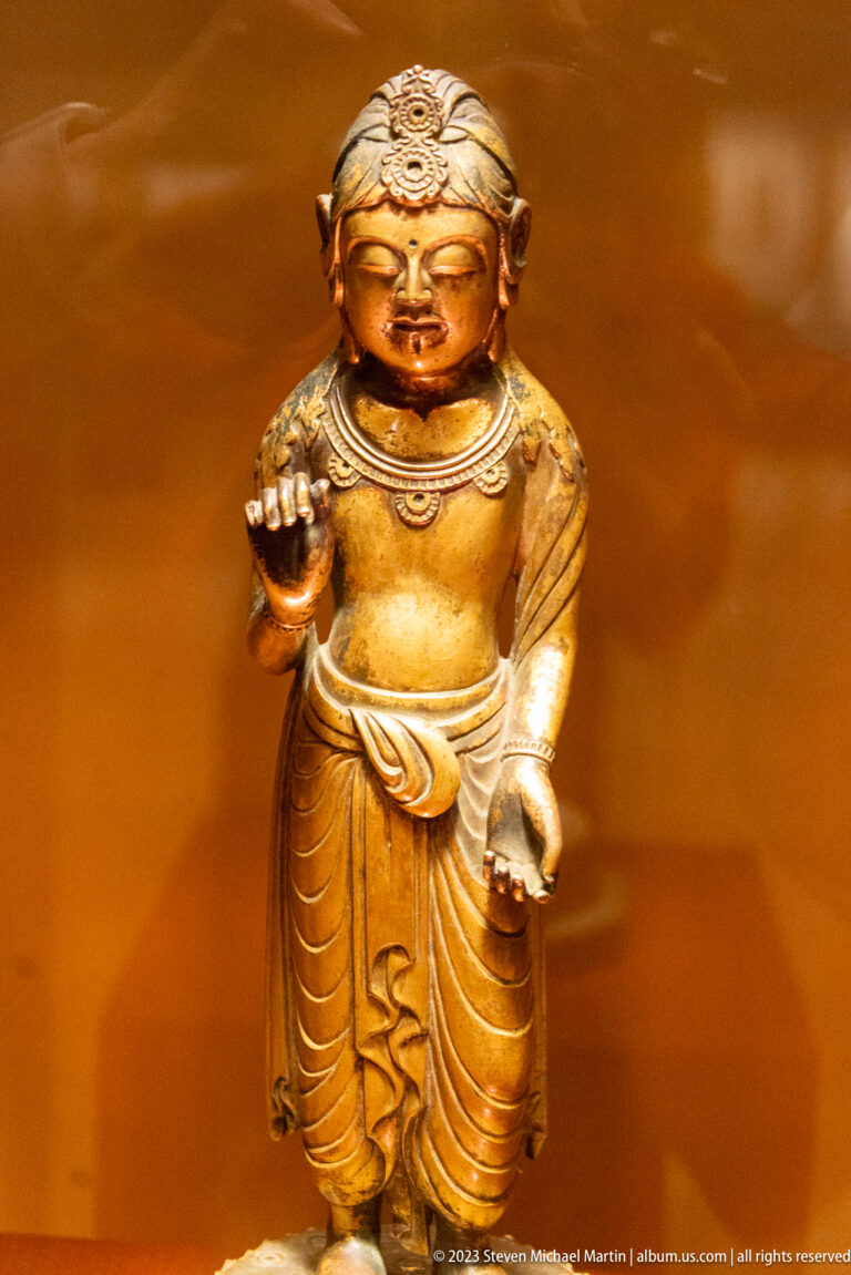Small golden Buddhist attendant with one raised hand and deeply folded drapery.