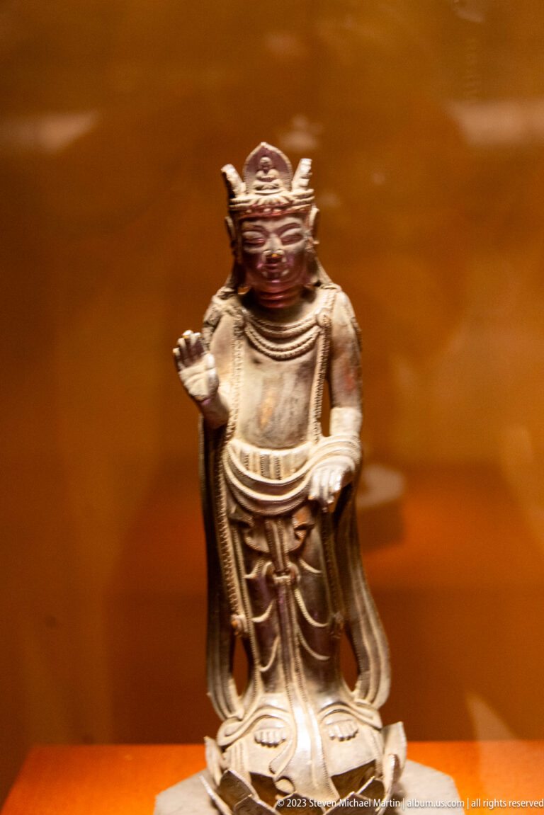 Small crowned bronze Buddhist figure on a lotus base with one palm raised.