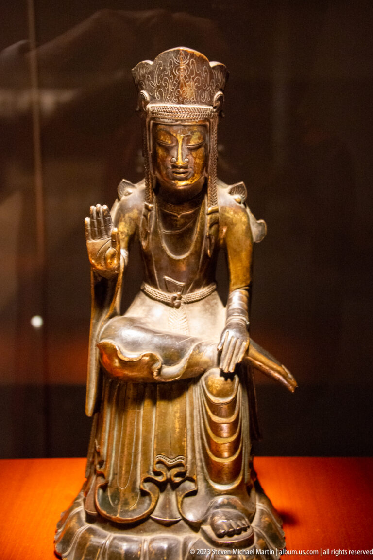 Seated crowned bronze Buddhist figure with one hand raised and a long narrow attribute behind the body.