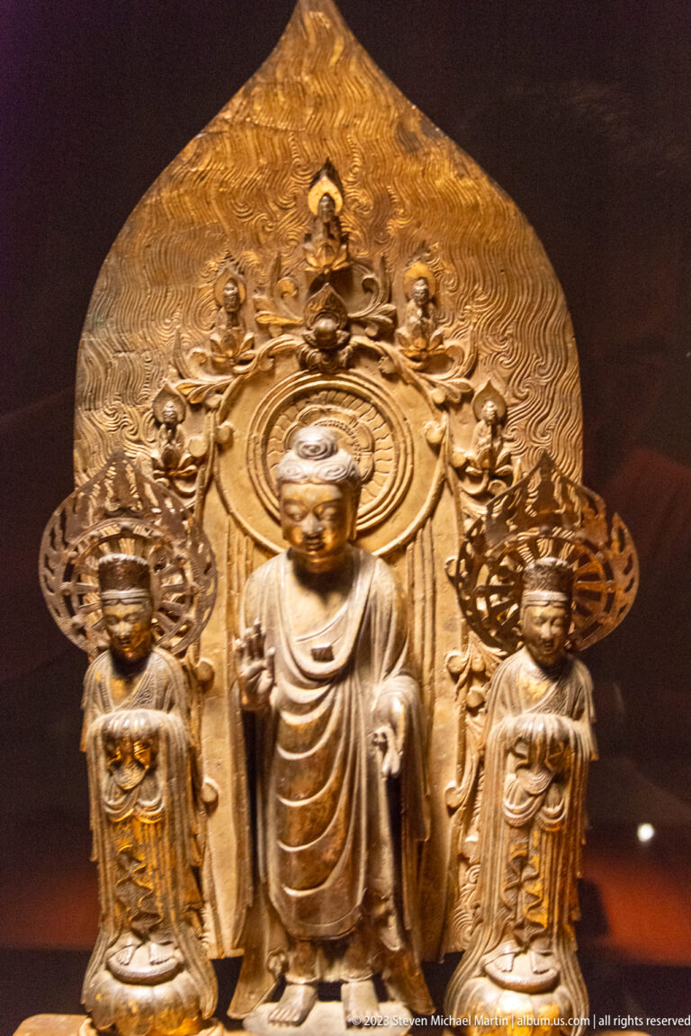 Bronze Buddhist triad with seated central Buddha, two standing attendants, and a large decorated mandorla.