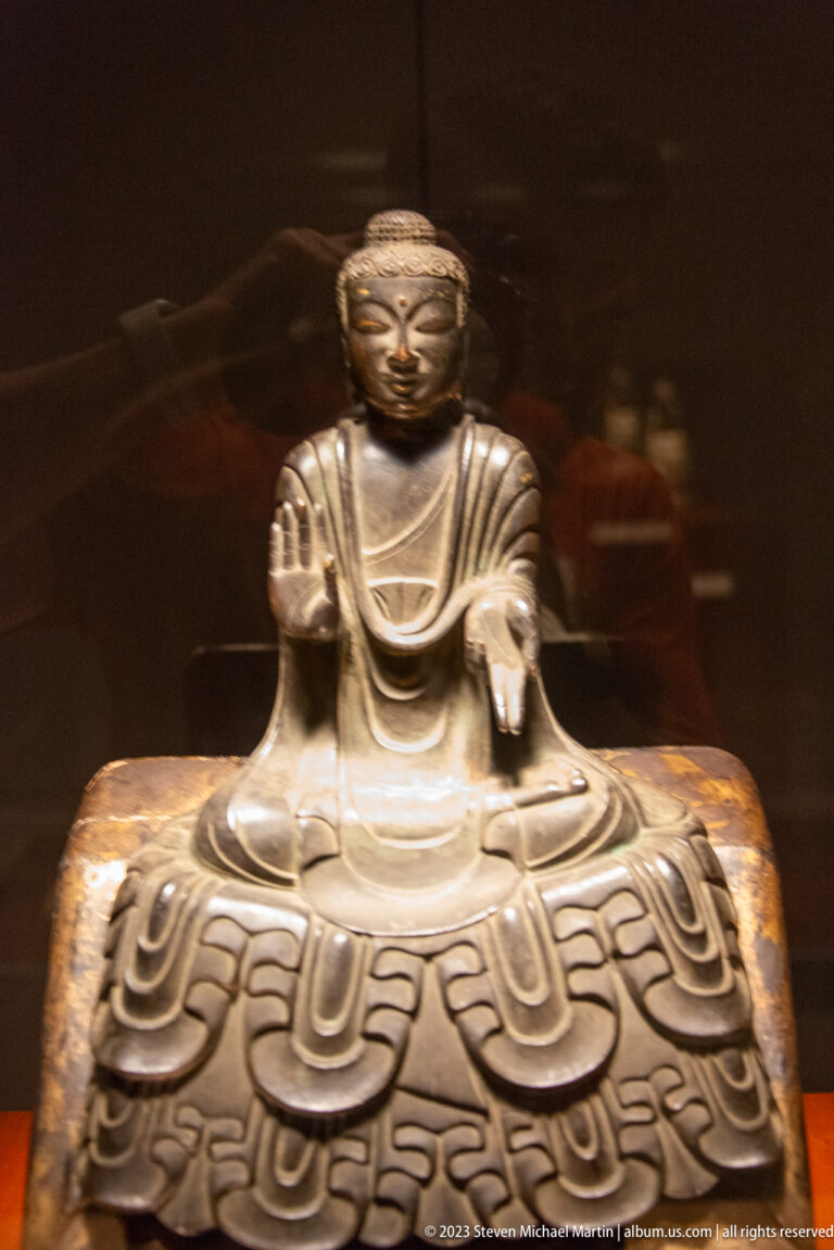 Small seated bronze Buddha with one hand raised on a broad pedestal with scrolling decoration.
